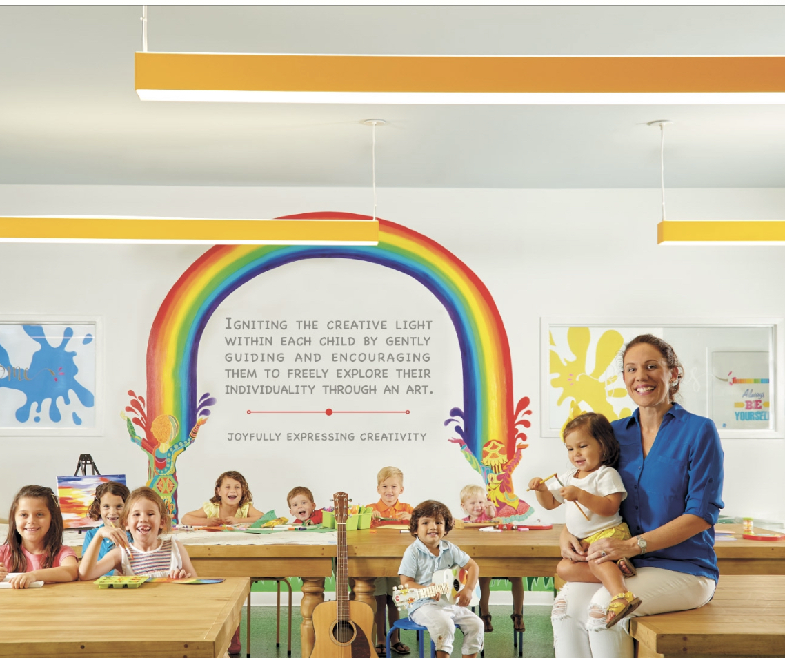 Children painting and playing instruments at a long wooden table beneath a hand-painted rainbow mural, with CJ Valle holding a toddler